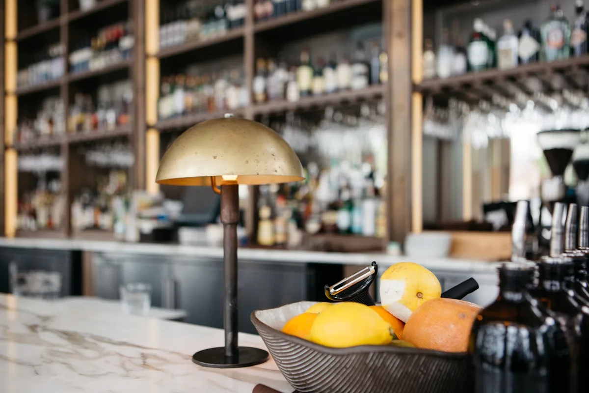 A brass dome lamp on a marble bar top, a bowl of lemons and oranges in front of a wood-framed bottle wall in soft focus, the kind of small set piece that anchors a room
