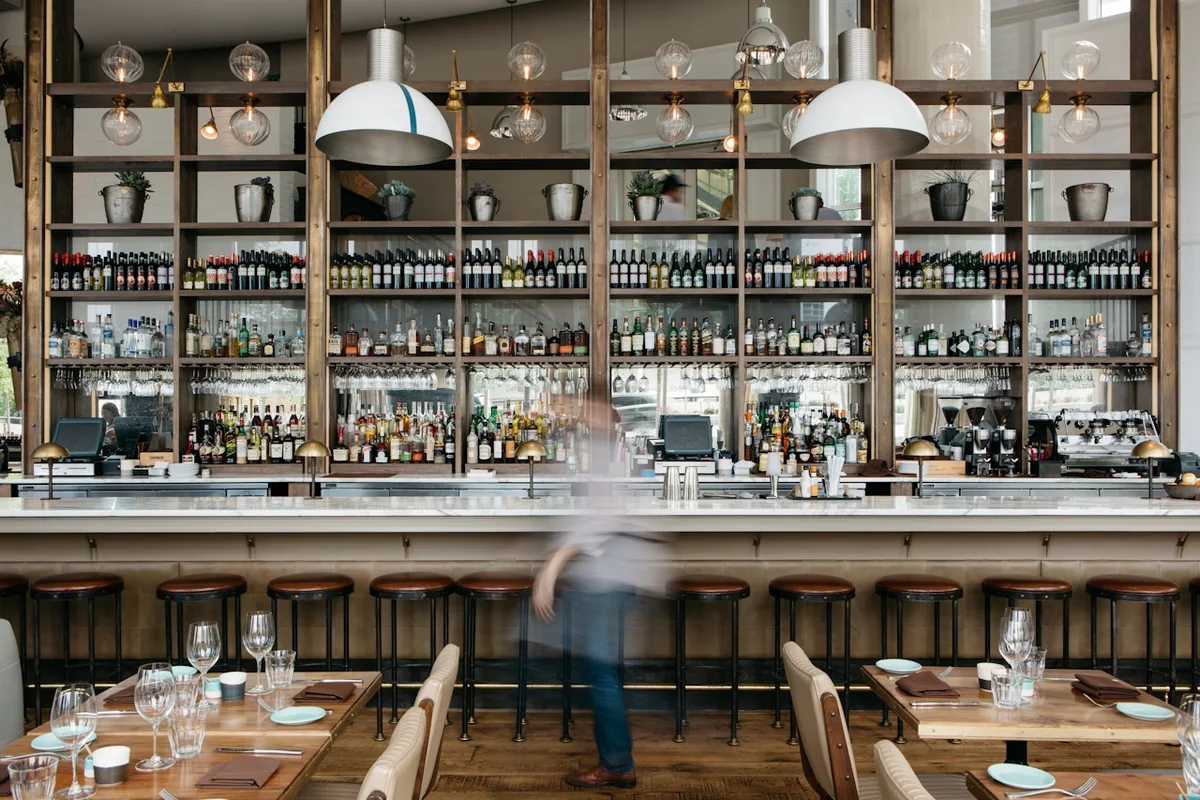 A wide editorial restaurant bar interior, wood-and-glass back wall lined with bottles, white pendant lights overhead, marble bar with stools, a figure walking past in soft motion blur