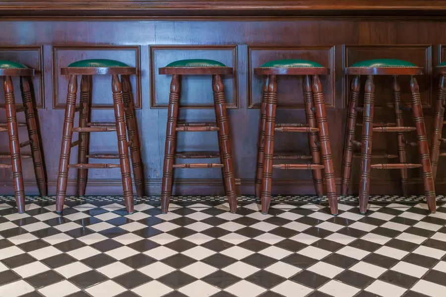 An empty row of turned-leg bar stools with green leather seats above a checkerboard tile floor